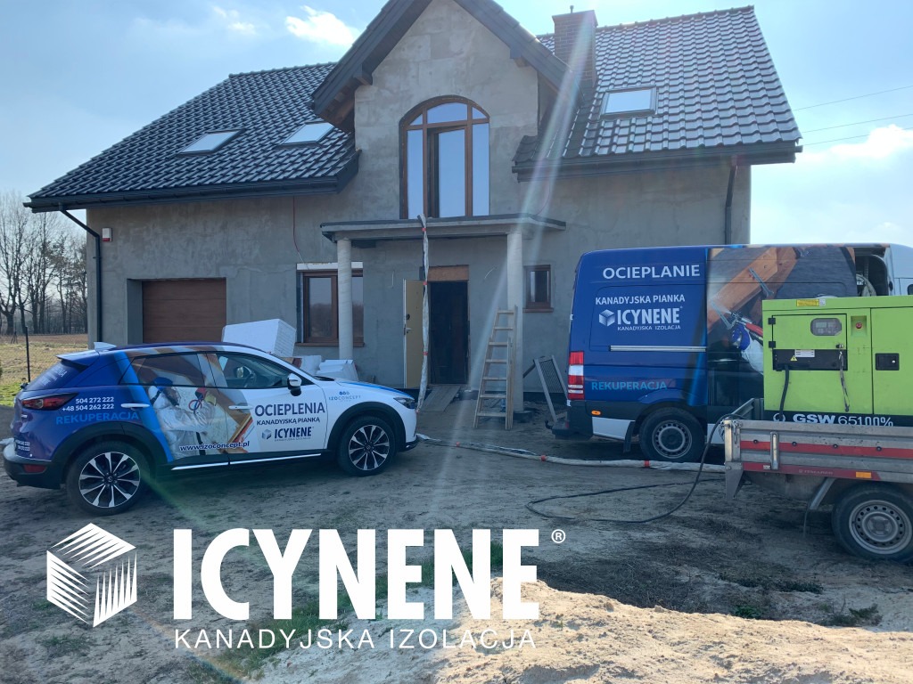 Dom w trakcie prac, z zaparkowanymi pojazdami firmy. Widoczny samochód osobowy i bus z logo firmy. Na przyczepie agregat prądotwórczy. Drabina oparta o ścianę budynku.