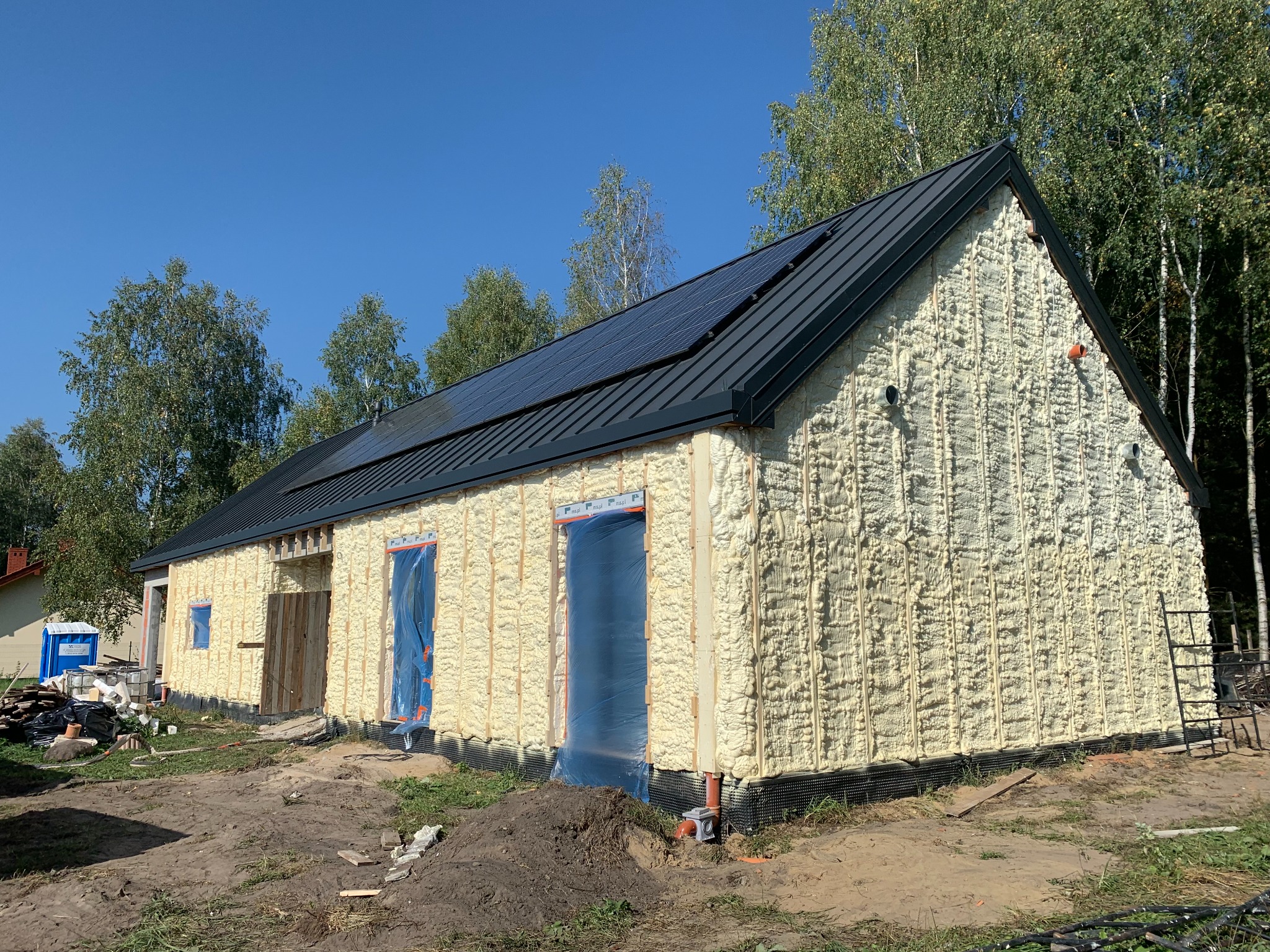 Budynek w trakcie izolacji: ściany pokryte pianą PUR, okna zabezpieczone folią. Widoczne panele słoneczne na dachu i fragment otoczenia z drzewami. Słoneczny dzień.