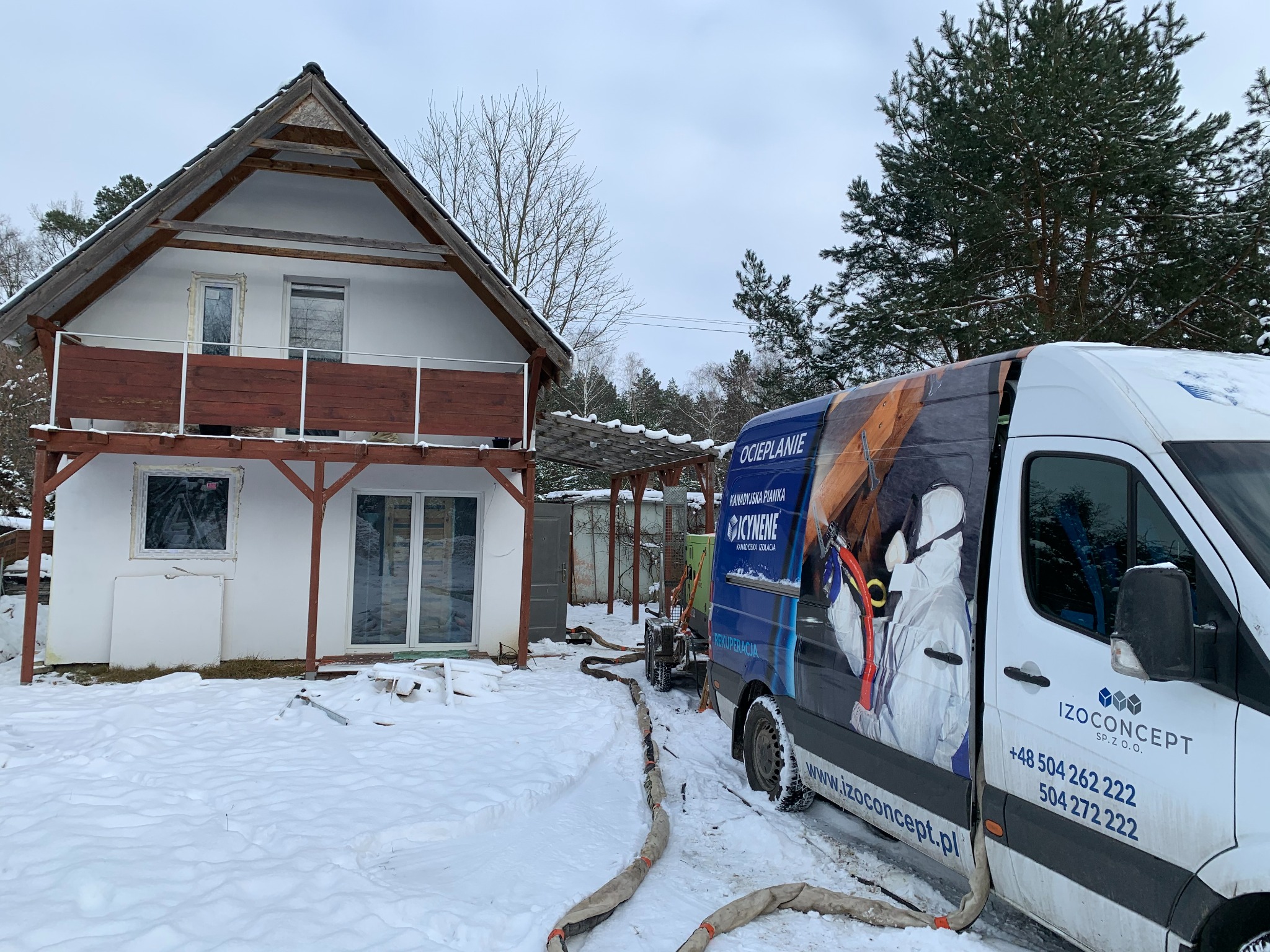 Ocieplanie pianką PUR: biały dom w zimowej scenerii, obok zaparkowany van z reklamą firmy IzoConcept i wężami do natrysku pianki. Widoczny etap przygotowania do ocieplenia.