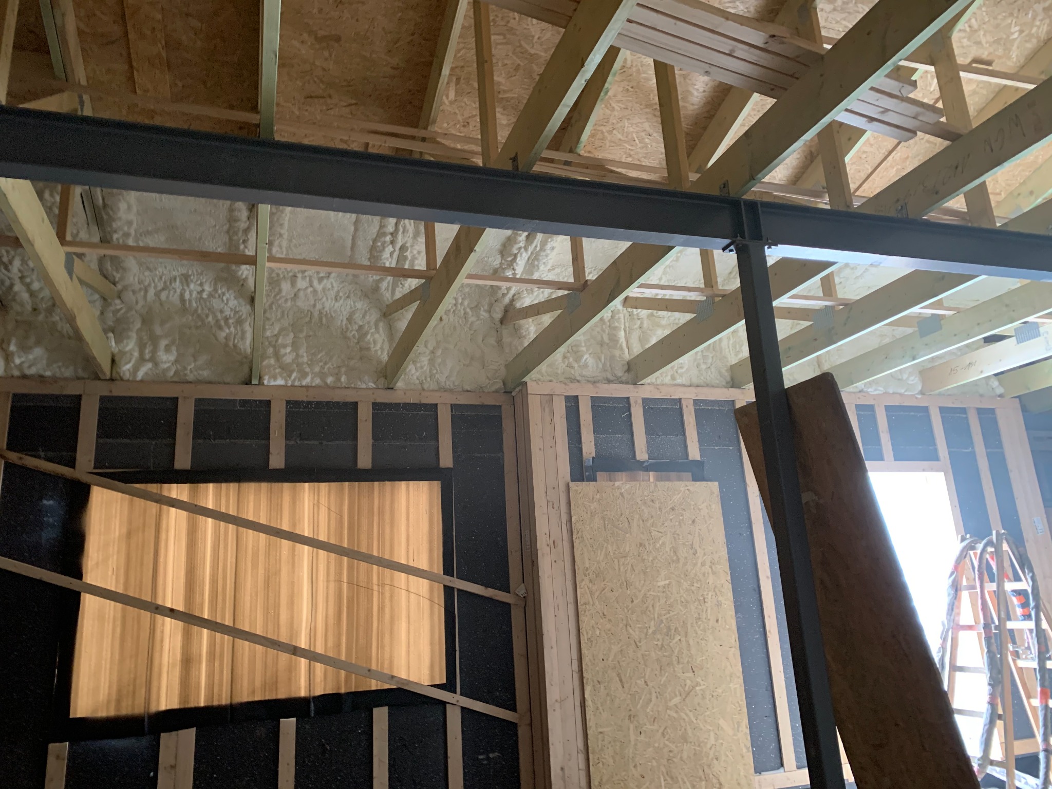 Wnętrze w trakcie prac: ściany z czarną izolacją i drewnianą konstrukcją, sufit z pianką izolacyjną, stalowa belka. Widoczna drabina i płyta OSB.