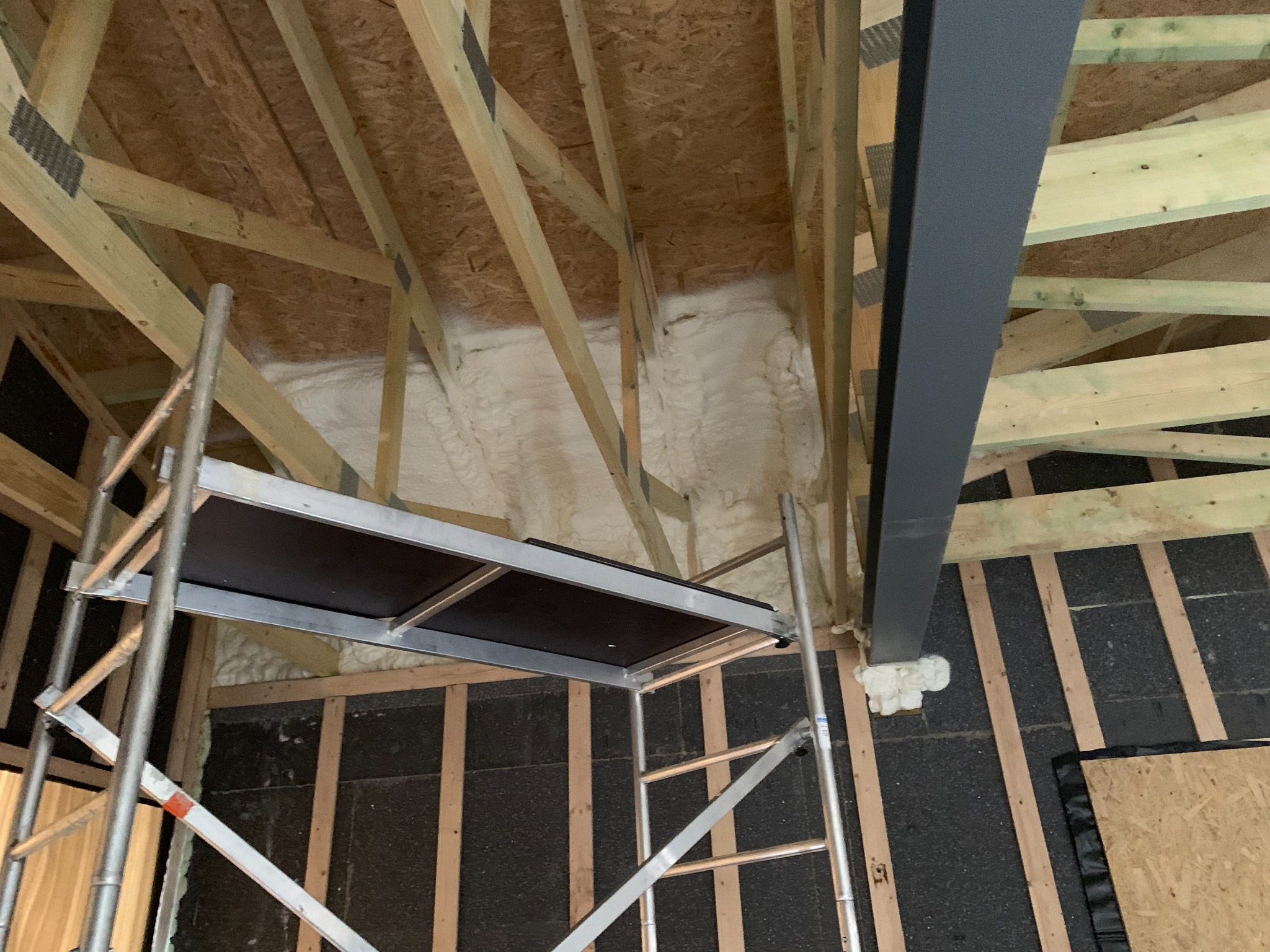 Skośny kadr poddasza z drewnianą konstrukcją, częściowo wypełnioną białą pianą izolacyjną. Widoczny fragment rusztowania i stalowy element konstrukcyjny.