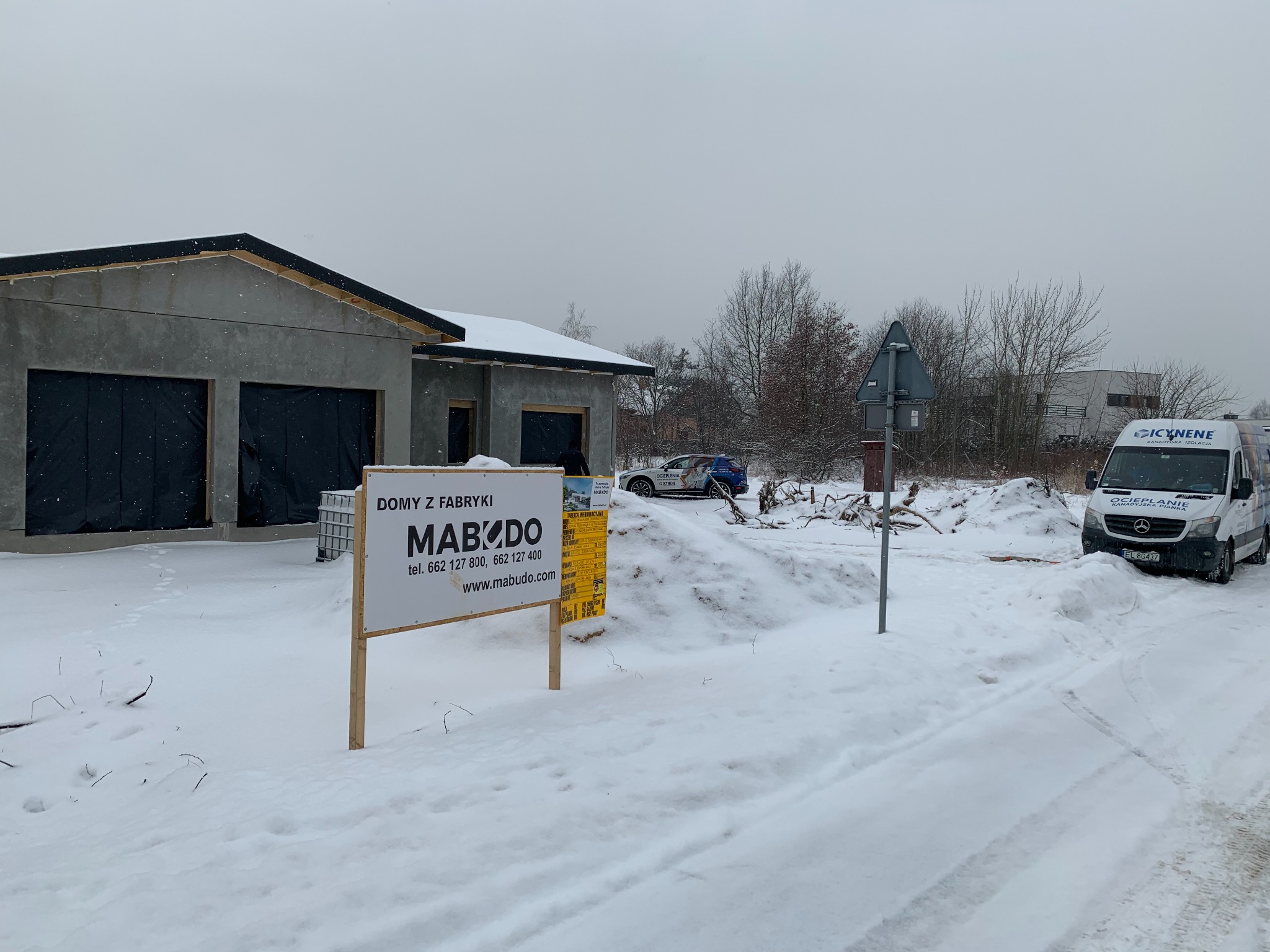 Surowy budynek w zimowej scenerii z tablicą informacyjną firmy budowlanej i samochodem z napisem 'Ocieplanie'. Widok na plac budowy w trakcie prac.