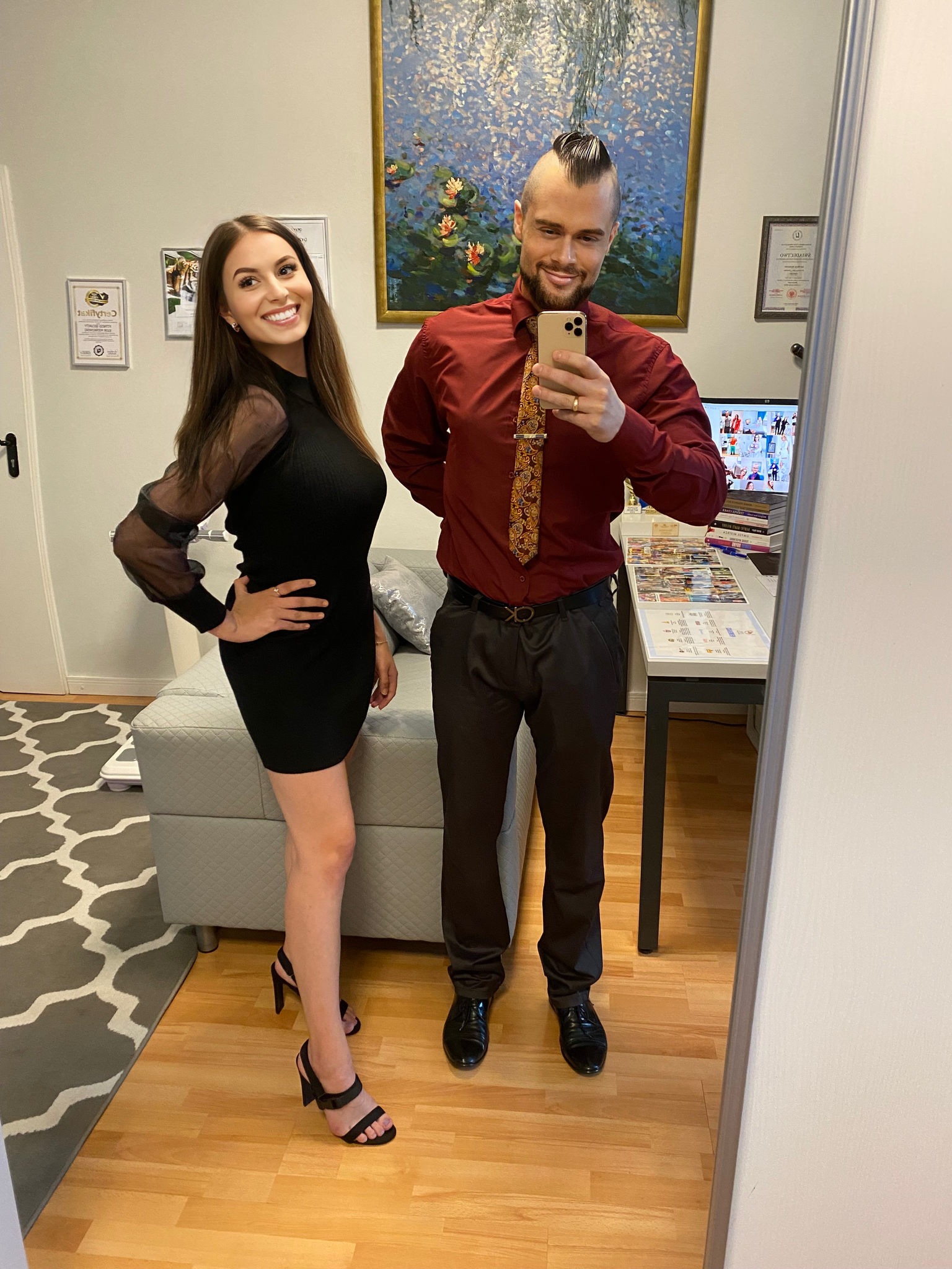 Para ubrana elegancko pozuje do selfie w lustrze, kobieta w czarnej sukience i szpilkach, mężczyzna w bordowej koszuli, spodniach i krawacie z wzorem.