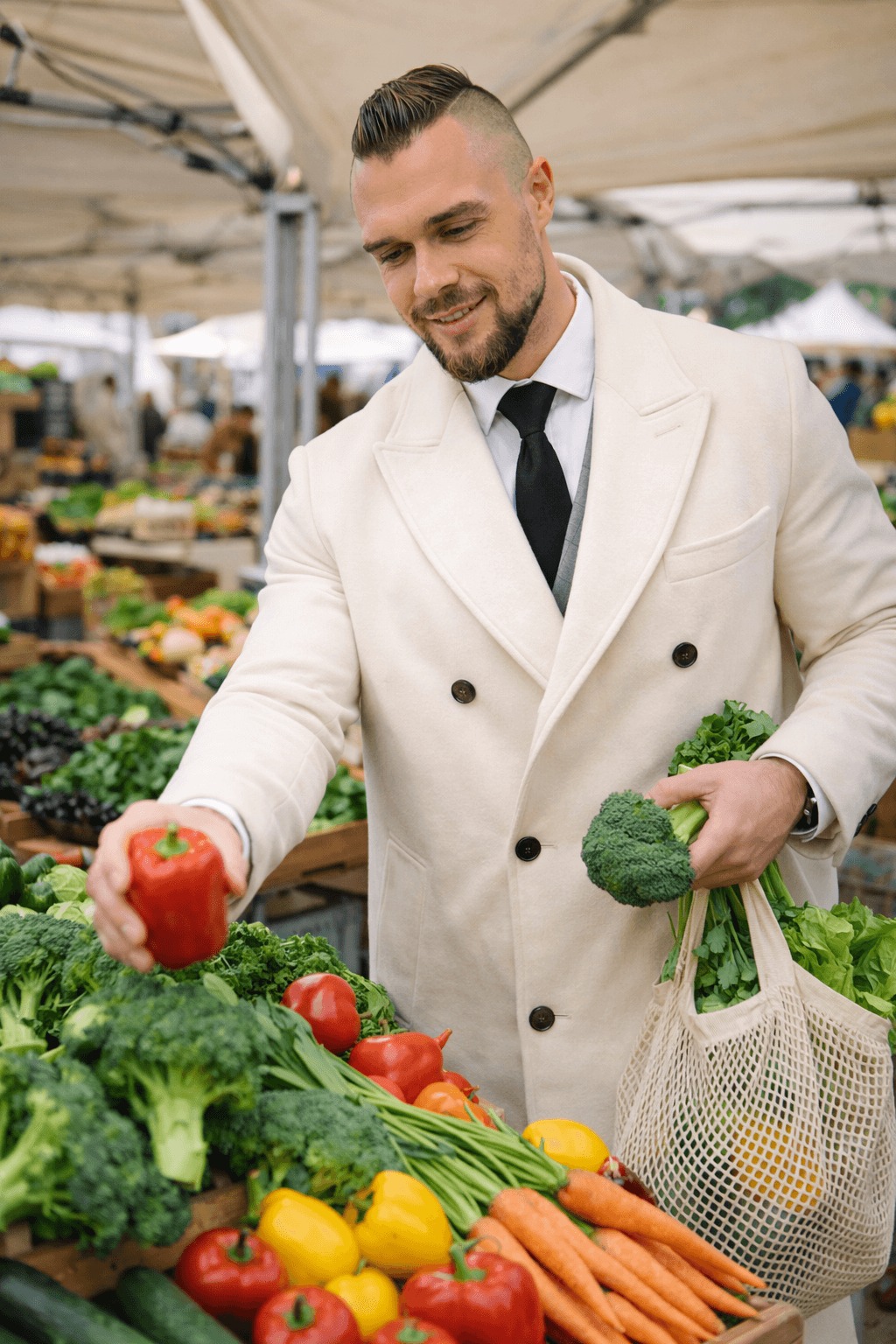 Mężczyzna w jasnym płaszczu wybiera czerwoną paprykę na straganie z warzywami: brokuły, marchew, pomidory i sałata w siatce.