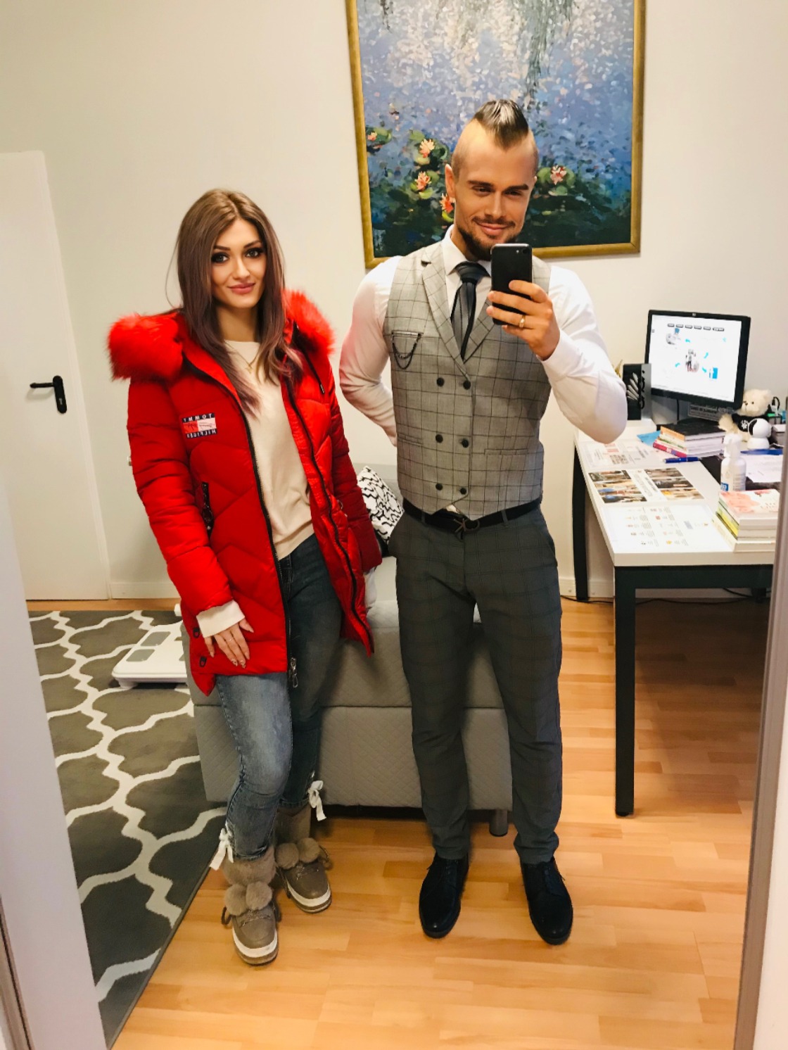 Para pozuje do selfie w gabinecie. Mężczyzna w kamizelce i krawacie trzyma telefon. Kobieta w czerwonej kurtce z futrem. W tle biurko z komputerem i obraz.