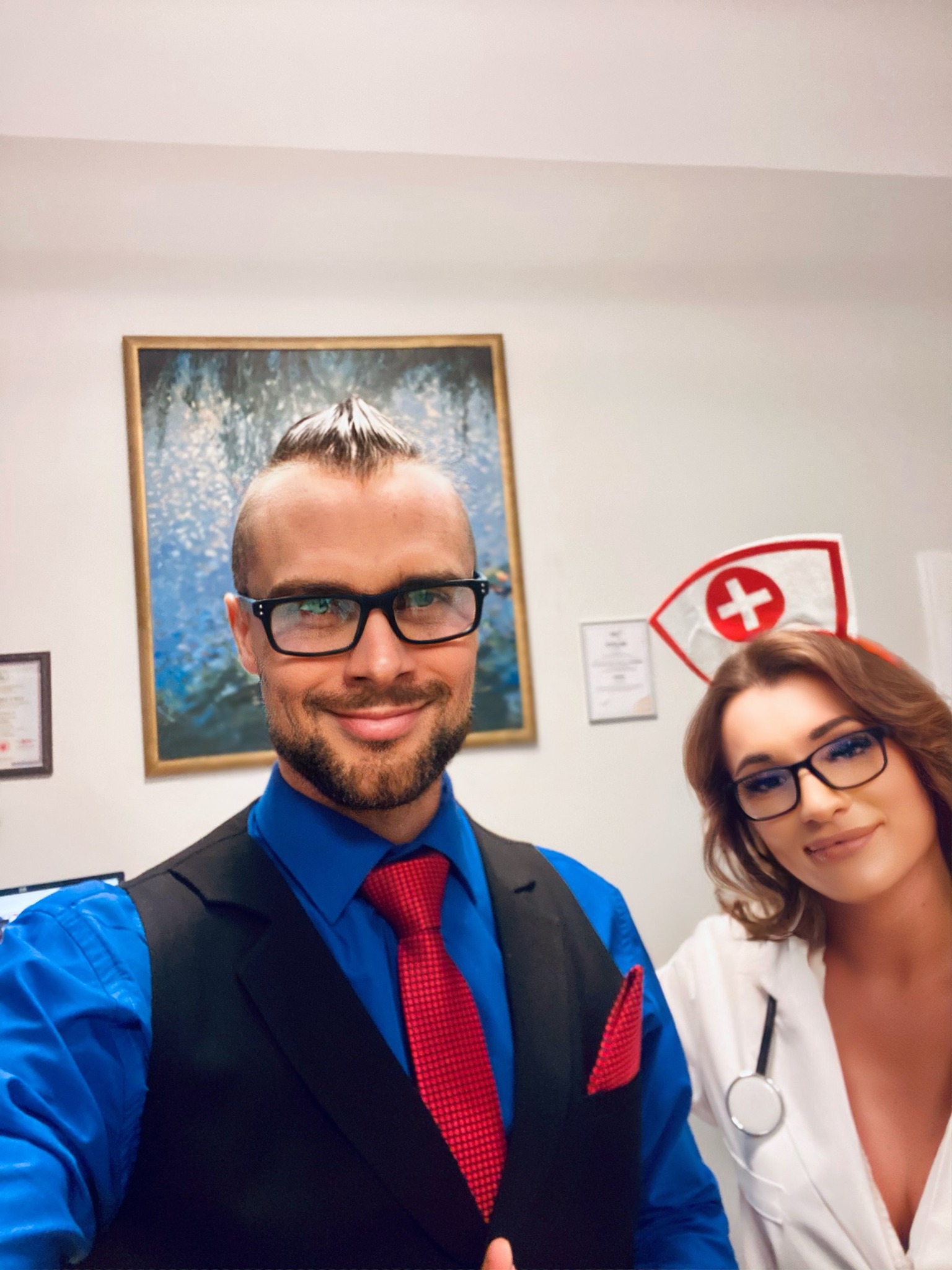 Portret dwojga uśmiechniętych pracowników medycznych: mężczyzna w niebieskiej koszuli, czarnej kamizelce i czerwonym krawacie oraz kobieta w stroju pielęgniarki ze stetoskopem, oboje w okularach...