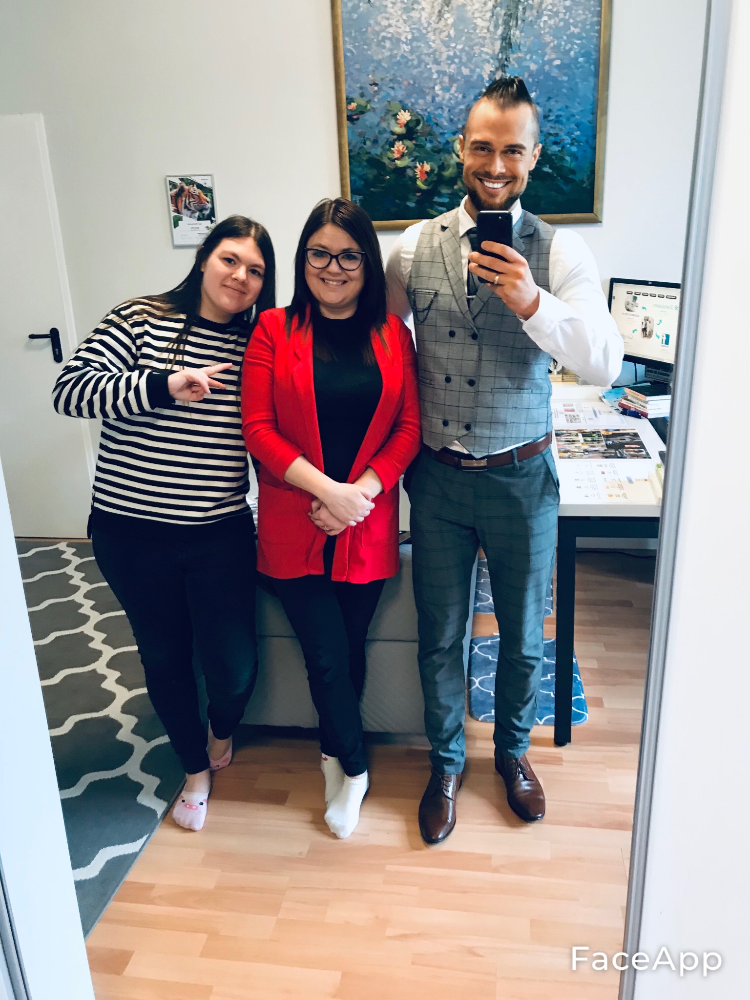Trzy osoby pozują do selfie w gabinecie, mężczyzna w eleganckiej kamizelce i spodniach w kratę trzyma telefon, na biurku widoczne materiały informacyjne, na ścianie obraz z liliami wodnymi.