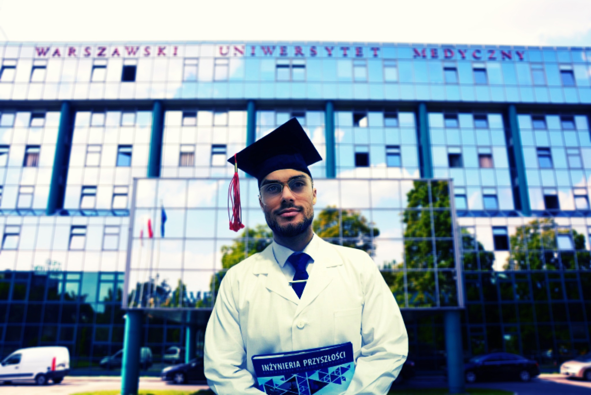 Absolwent w todze i okularach, trzymający książkę 'Inżynieria Przyszłości' na tle budynku Warszawskiego Uniwersytetu Medycznego.