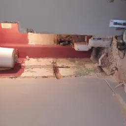 Wnętrze łazienki w trakcie remontu, widoczne ślady po skuciu płytek, odsłonięte rury, bojler na ścianie, szpachla na ścianie, narzędzia budowlane i worki z zaprawą w tle.