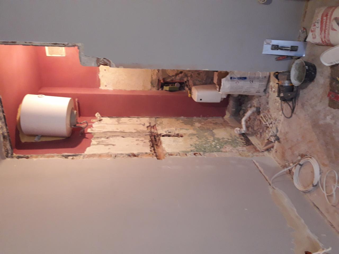 Wnętrze łazienki w trakcie remontu, widoczne ślady po skuciu płytek, odsłonięte rury, bojler na ścianie, szpachla na ścianie, narzędzia budowlane i worki z zaprawą w tle.