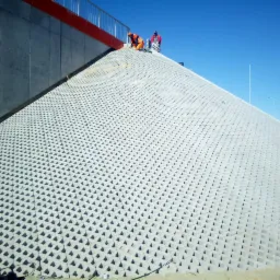 Skarpa wzmocniona ażurowymi płytami betonowymi, widoczni dwaj pracownicy w odblaskowych ubraniach na górze nasypu, niebieskie niebo w tle.