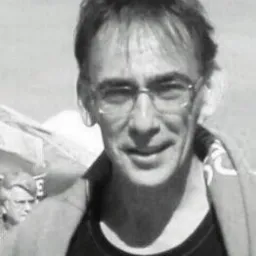 Czarno-białe zbliżenie uśmiechniętego mężczyzny w okularach, zarys postaci w tle, na zewnątrz. Portret w swobodnej atmosferze.