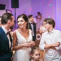 Uśmiechnięta panna młoda w koronkowej sukni rozmawia z panem młodym w granatowym garniturze, w tle goście weselni i dekoracje świetlne.