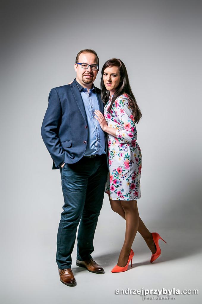 Portret pary: mężczyzna w niebieskiej marynarce i okularach, kobieta w kwiecistej sukience i pomarańczowych szpilkach, na jasnoszarym tle.