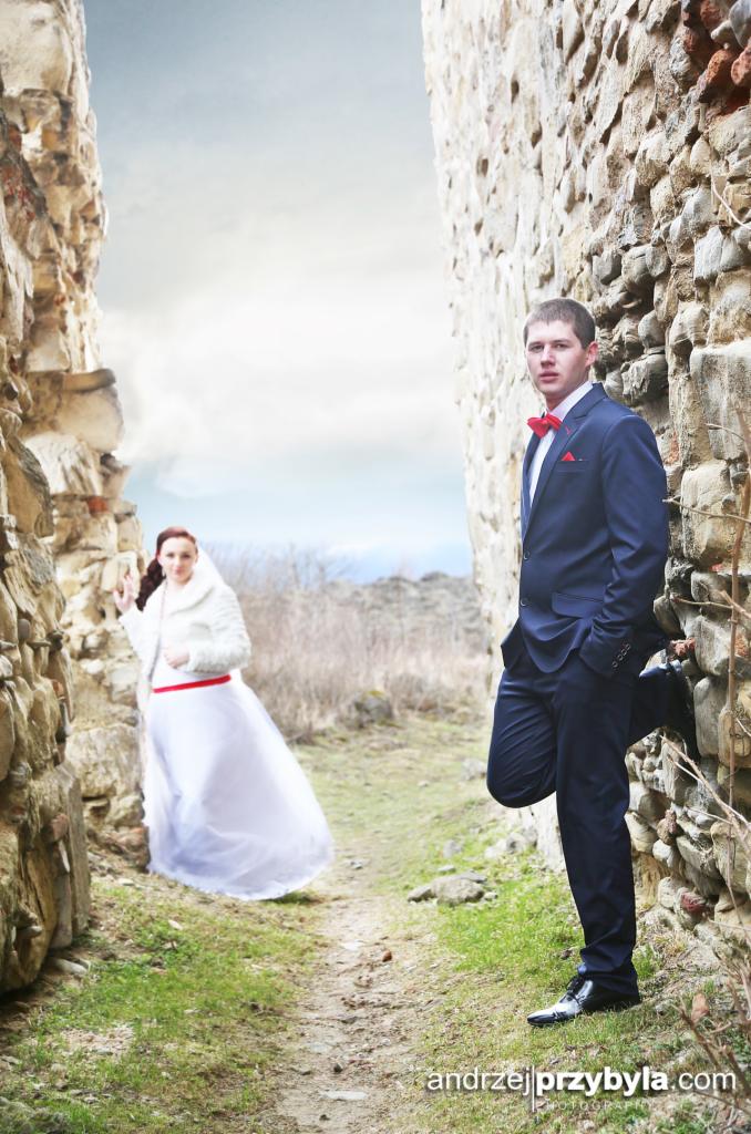 Para młoda w ruinach zamku: Pan młody w granatowym garniturze z czerwoną muchą opiera się o kamienną ścianę, panna młoda w białej sukni z czerwonym paskiem w tle, w otoczeniu kamiennych murów...