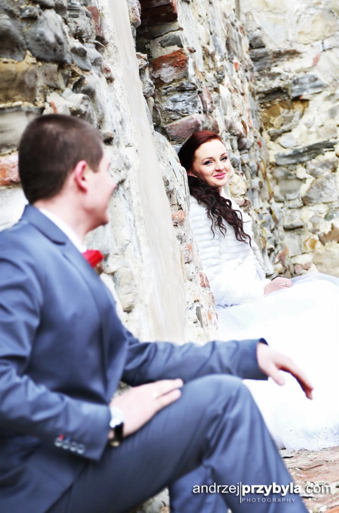 Para młoda siedzi na kamiennych schodach, opierając się o starą, ceglaną ścianę; panna młoda w białej sukni i futrzanym bolerku uśmiecha się, patrząc na pana młodego w granatowym garniturze...