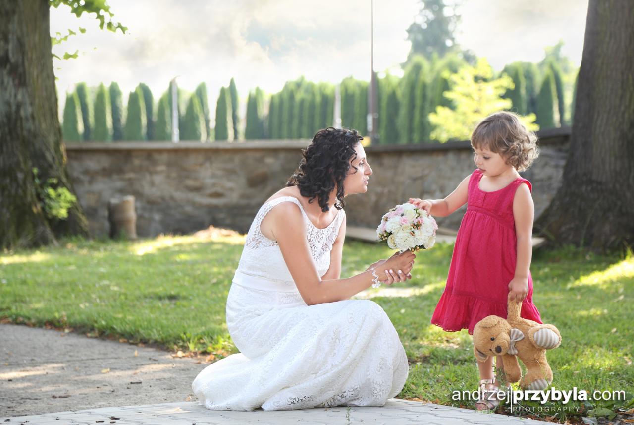 Panna młoda w koronkowej sukni klęczy na ścieżce, podając bukiet kwiatów małej dziewczynce w różowej sukience trzymającej pluszowego misia, w tle zielony żywopłot.