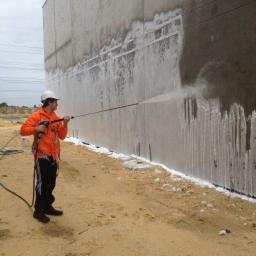 CleanBlast - innowacyjne czyszczenie powierzchni - Pracownik w kasku i pomarańczowej kurtce czyści betonową ścianę budynku myjką ciśnieniową, widoczne zacieki i piana na ziemi.
