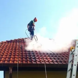 Pracownik w kasku i uprzęży czyści dach z czerwonej dachówki za pomocą myjki ciśnieniowej, obok drabina oparta o dach, w tle błękitne niebo.