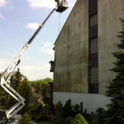 Pracownik w koszu podnośnika teleskopowego czyści elewację budynku z widocznymi zabrudzeniami i zaciekami, zieleń wokół budynku.