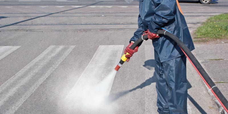 Pracownik w niebieskim kombinezonie i czerwonych rękawicach czyści przejście dla pieszych za pomocą myjki ciśnieniowej, strumień wody usuwa zabrudzenia z białych pasów zebry.