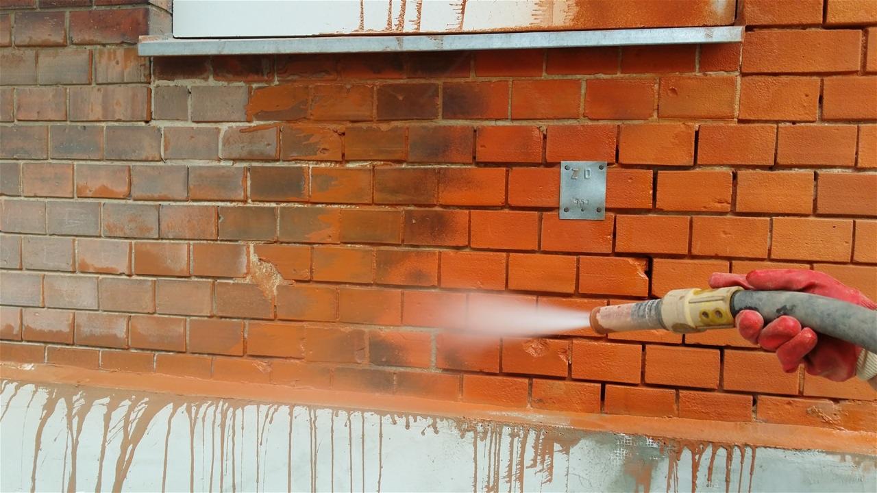 Ceglana ściana czyszczona strumieniem wody pod ciśnieniem, widoczne różnice w kolorze cegieł przed i po czyszczeniu, gumowe rękawice ochronne.
