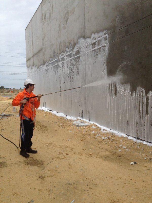 Pracownik w kasku i pomarańczowej kurtce czyści betonową ścianę budynku myjką ciśnieniową, widoczne zacieki i piana na ziemi.