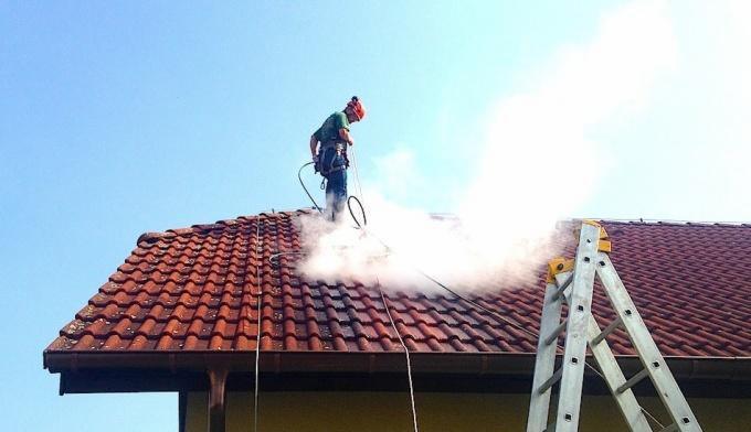 Pracownik w kasku i uprzęży czyści dach z czerwonej dachówki za pomocą myjki ciśnieniowej, obok drabina oparta o dach, w tle błękitne niebo.