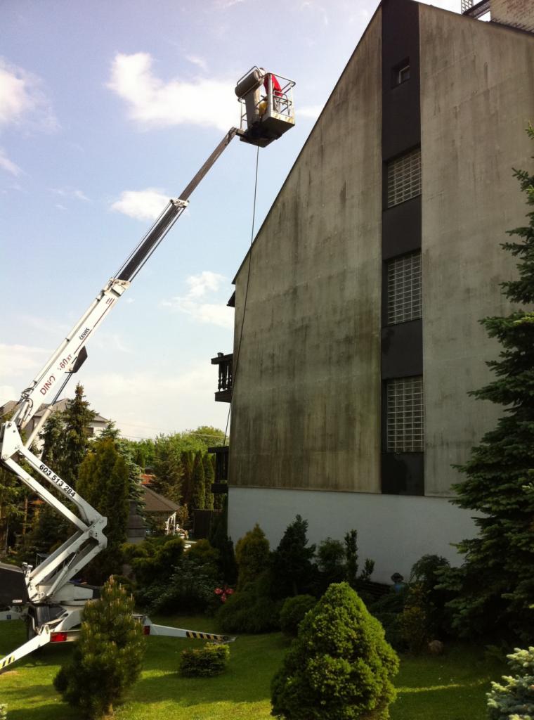 Pracownik w koszu podnośnika teleskopowego czyści elewację budynku z widocznymi zabrudzeniami i zaciekami, zieleń wokół budynku.