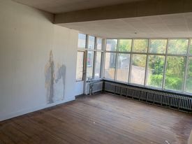 Puste pomieszczenie w trakcie przygotowań do remontu, widoczne ślady po wcześniejszych pracach na ścianie, stare drewniane podłogi i grzejniki pod oknami.