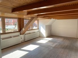 Puste, jasne poddasze z drewnianymi belkami stropowymi i skośnym słupem. Widoczne dwa okna z grzejnikami pod nimi oraz fragment schodów. Podłoga pokryta panelami w odcieniach szarości i beżu.