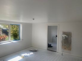 Białe, świeżo pomalowane wnętrze pokoju z oknem i otwartymi drzwiami. Na podłodze widoczny talerz malarski, a przy ścianie wałek oparty o szpachlowaną powierzchnię.