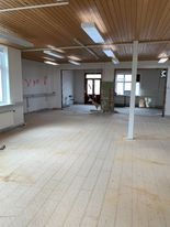 Puste, jasne pomieszczenie w trakcie remontu. Widoczne surowe drewniane podłogi, białe ściany z pozostałościami po instalacjach, drewniany sufit z lampami i filarem pośrodku. W głębi widoczne...