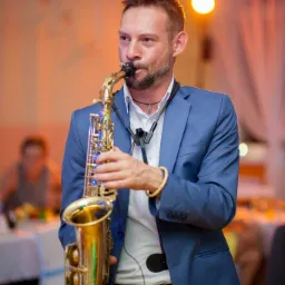 Saksofonista w niebieskim garniturze grający na złotym saksofonie podczas wydarzenia z oświetleniem scenicznym.