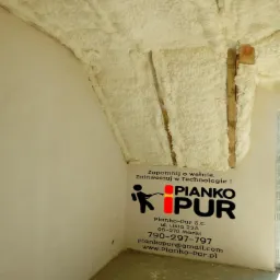 Poddasze ocieplone białą pianką poliuretanową, widoczne drewniane elementy konstrukcyjne, na ścianie naklejka reklamowa firmy wykonującej ocieplenie.