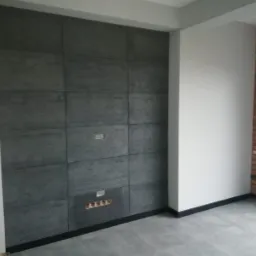Ściana wewnętrzna wykończona szarymi panelami imitującymi beton, z widocznymi puszkami elektrycznymi i fragmentem ściany z cegły dekoracyjnej.