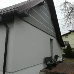 Świeżo otynkowana, szara elewacja domu z grafitowymi pasami, ciemnobrązowe rynny, widoczna ławka i fragment posesji w pochmurny dzień.