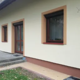 Elewacja domu jednorodzinnego w jasnych kolorach z brązowymi obramowaniami okien i drzwi; widoczny fragment kostki brukowej przed wejściem.