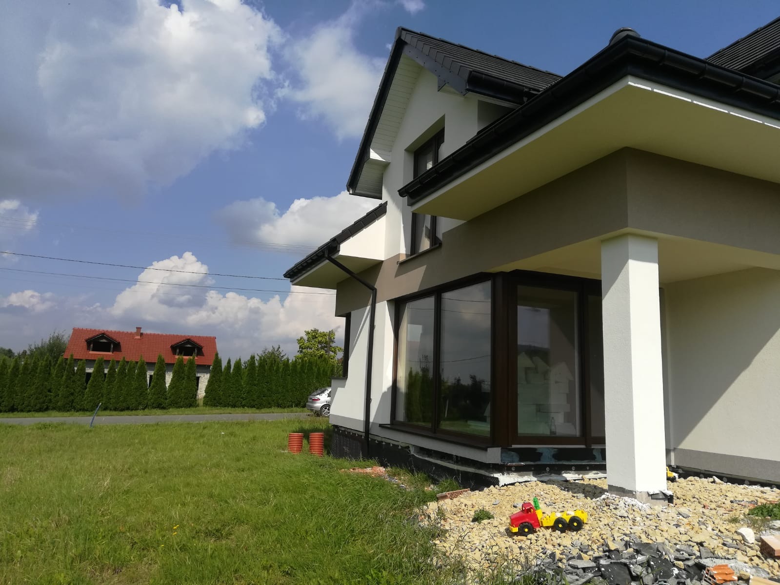 Fragment nowo wybudowanego domu z dużymi oknami, widoczne niedokończone prace ziemne i zabawka dziecka na gruzie.
