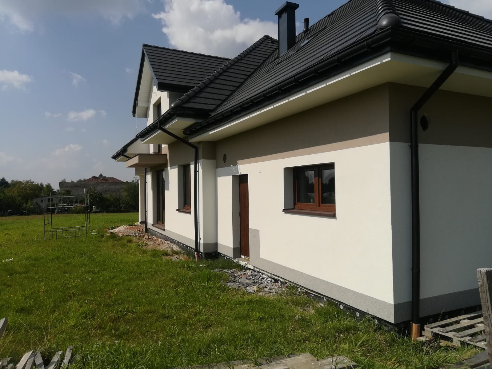 Nowoczesny dom jednorodzinny w Żywcu z dwukolorową elewacją, czarną dachówką i orynnowaniem, widoczny z boku na tle zielonej trawy i błękitnego nieba z chmurami.