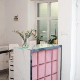 Recepcja w salonie kosmetycznym z marmurowym blatem, różowym panelem tapicerowanym i bukietem różowych goździków w wazonie.