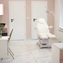 Jasne wnętrze gabinetu kosmetycznego z białym fotelem zabiegowym, umywalką z czarną misą i marmurową posadzką.