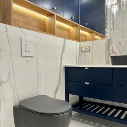 Archi-Ann Adam Skrzypczyński - Nowoczesna łazienka z szarą podwieszaną toaletą, szafkami w kolorze granatowym z złotymi uchwytami, marmurowymi płytkami i oświetleniem LED w drewnianych półkach.