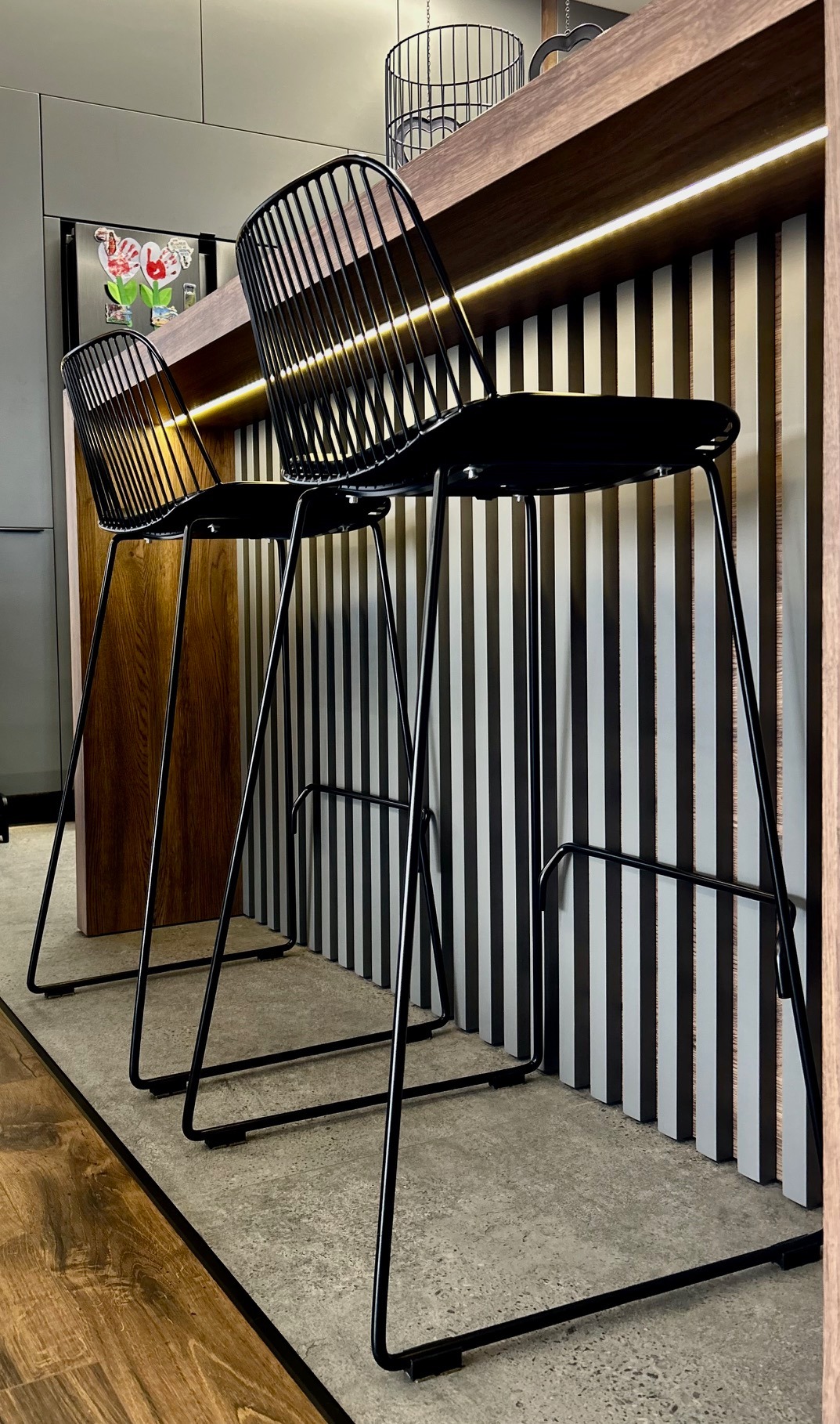 Dwa czarne, metalowe hokery barowe z oparciami, ustawione przy blacie z podświetleniem LED, na tle ściany z pionowymi, czarno-białymi panelami. Na lodówce widoczne rysunki dziecięce.