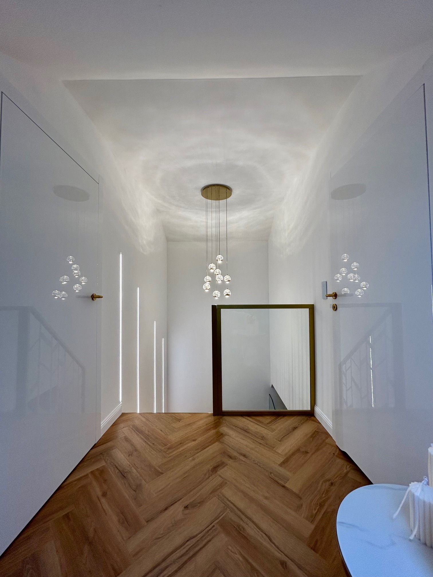 Nowoczesny korytarz z parkietem w jodełkę, białymi ścianami z ukrytymi drzwiami i lustrem w złotej ramie, oświetlony wiszącą lampą z kulistymi kloszami.