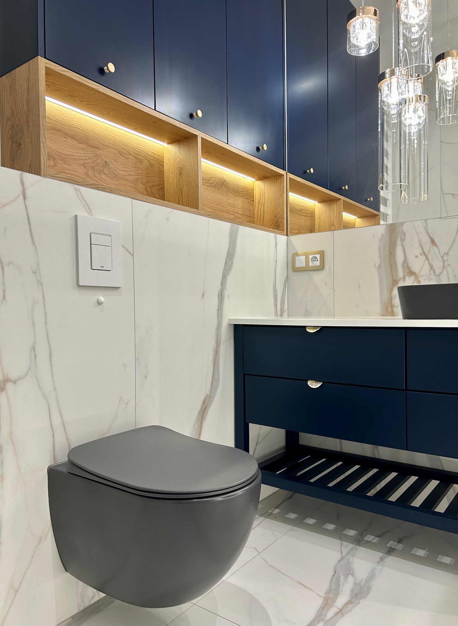 Nowoczesna łazienka z szarą podwieszaną toaletą, szafkami w kolorze granatowym z złotymi uchwytami, marmurowymi płytkami i oświetleniem LED w drewnianych półkach.
