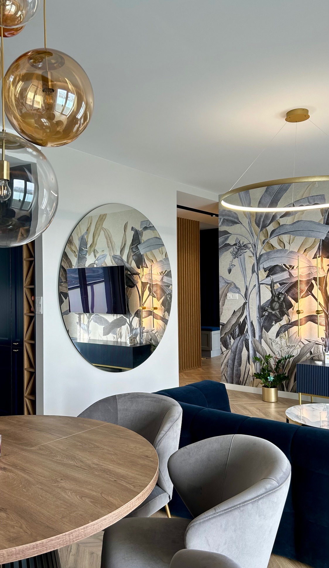Nowoczesne wnętrze: okrągły stół z szarymi krzesłami, lustro z wbudowanym telewizorem na tle tapety z motywem tropikalnych liści, designerskie lampy wiszące.