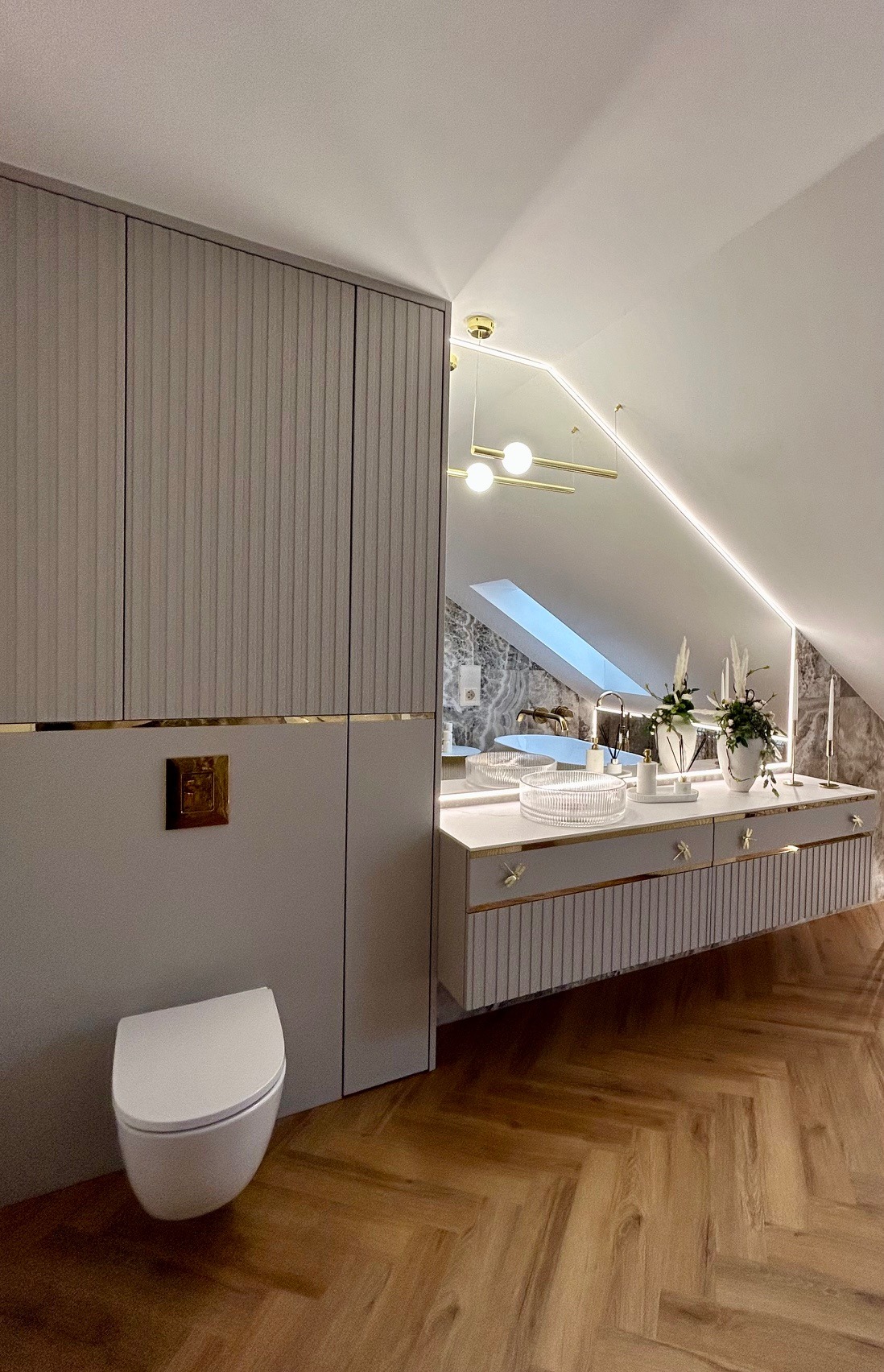 Elegancka łazienka na poddaszu ze skośnym sufitem, z zabudowaną szafą, wiszącą toaletą, umywalką wpuszczoną w blat z marmuru, złotymi akcentami i oświetleniem LED.