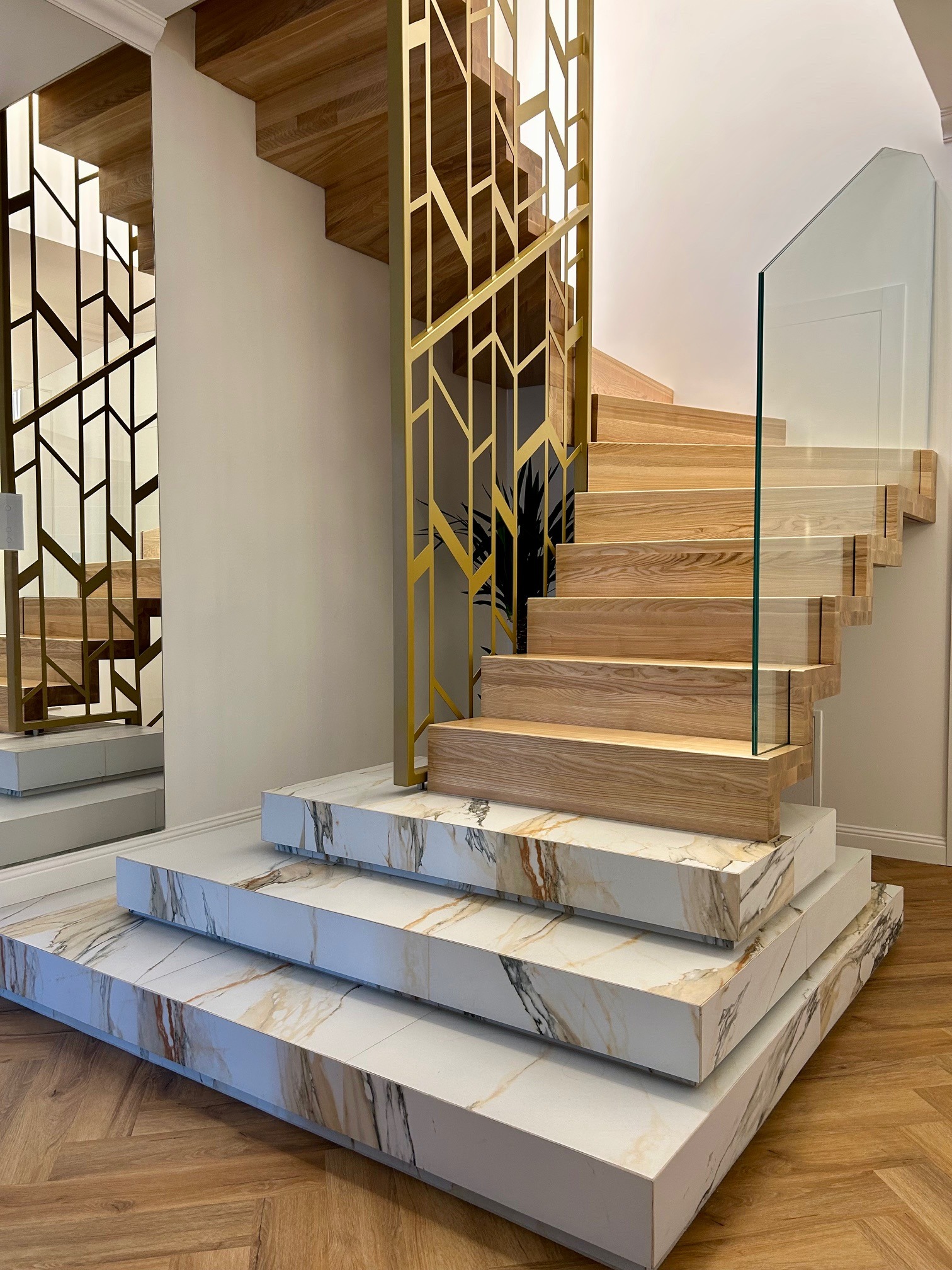 Nowoczesne schody z drewnianymi stopniami i marmurowymi podstopnicami, zabezpieczone szklaną balustradą i złotą, geometryczną konstrukcją, odbijające się w lustrze.
