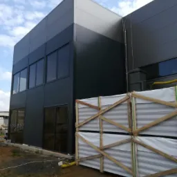 Nowoczesny budynek przemysłowy z elewacją z paneli warstwowych w odcieniach szarości, z oknami i przygotowanymi do montażu panelami w drewnianych ramach na pierwszym planie.